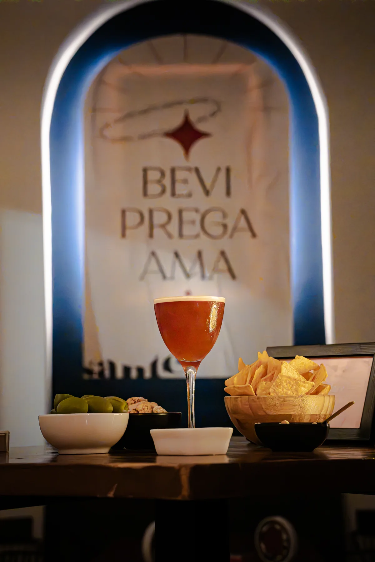 Cocktail e stuzzichini davanti alla scritta Bevi Prega Ama di Santé